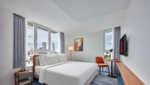 Ein Hotelzimmer mit einem großen Bett, einem Schreibtisch, einem Sessel, einem Fernseher und Blick auf die Stadt.