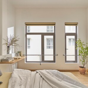 Une chambre à coucher avec un lit, une table de chevet avec des livres et un vase, deux fenêtres avec des stores et une plante en pot.