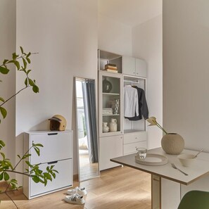 Une chambre moderne et minimaliste, dotée d’une armoire intégrée, d’un petit réfrigérateur, d’une table à manger agrémentée d’un vase et d’un verre, et d’une paire de chaussures posées sur le sol.