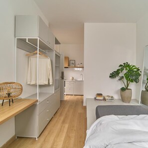 Een moderne slaapkamer met een bed, een bureau, een kledingkast, een kamerplantje en een kleine kitchenette.