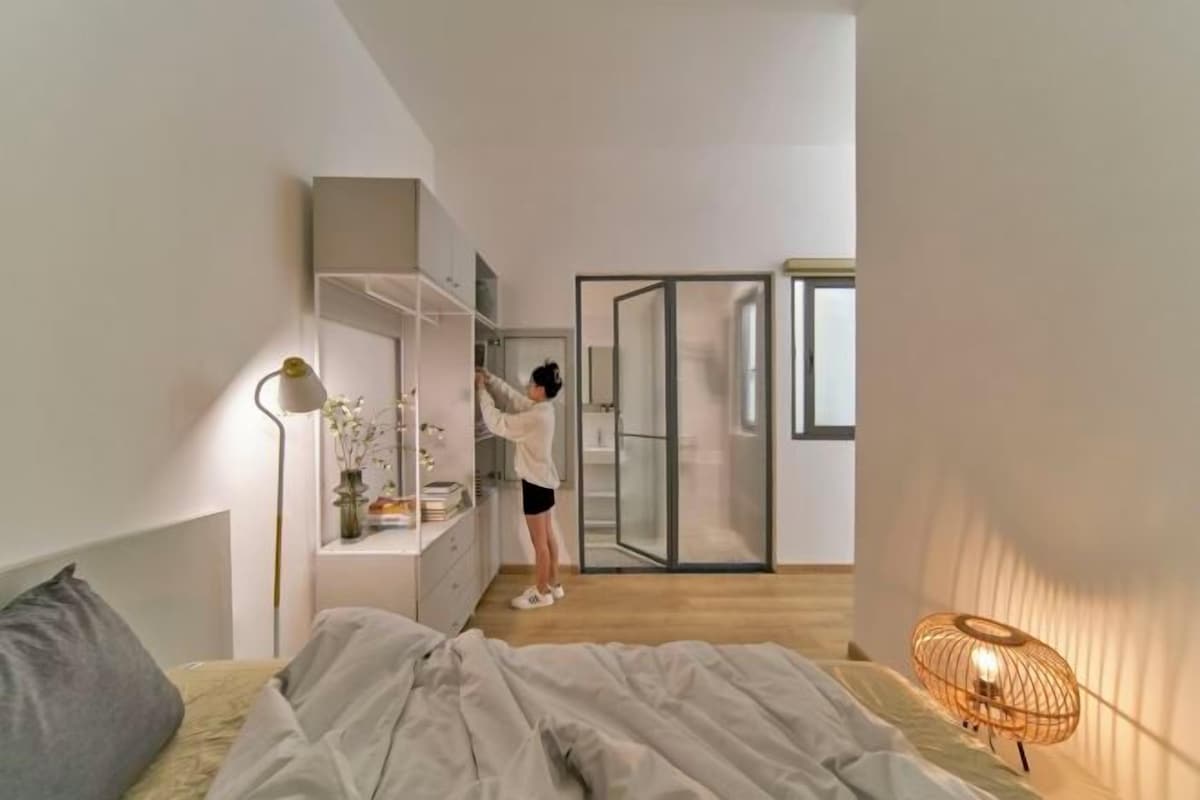 Een moderne slaapkamer met een bed, nachtkastje, lamp en een persoon die een plank opruimt.