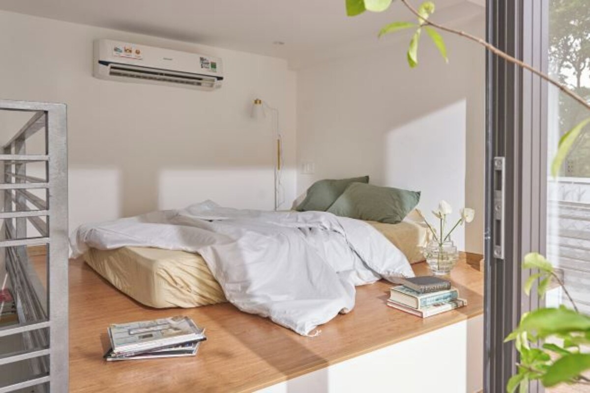 Une chambre à coucher avec un lit, une table de chevet avec des livres et un vase, une fenêtre donnant sur un beau paysage et un appareil de climatisation.