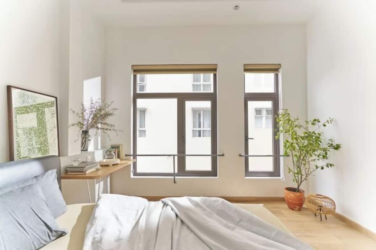 Une chambre à coucher avec un lit, deux fenêtres, une table de chevet, une plante et un tableau encadré au mur.