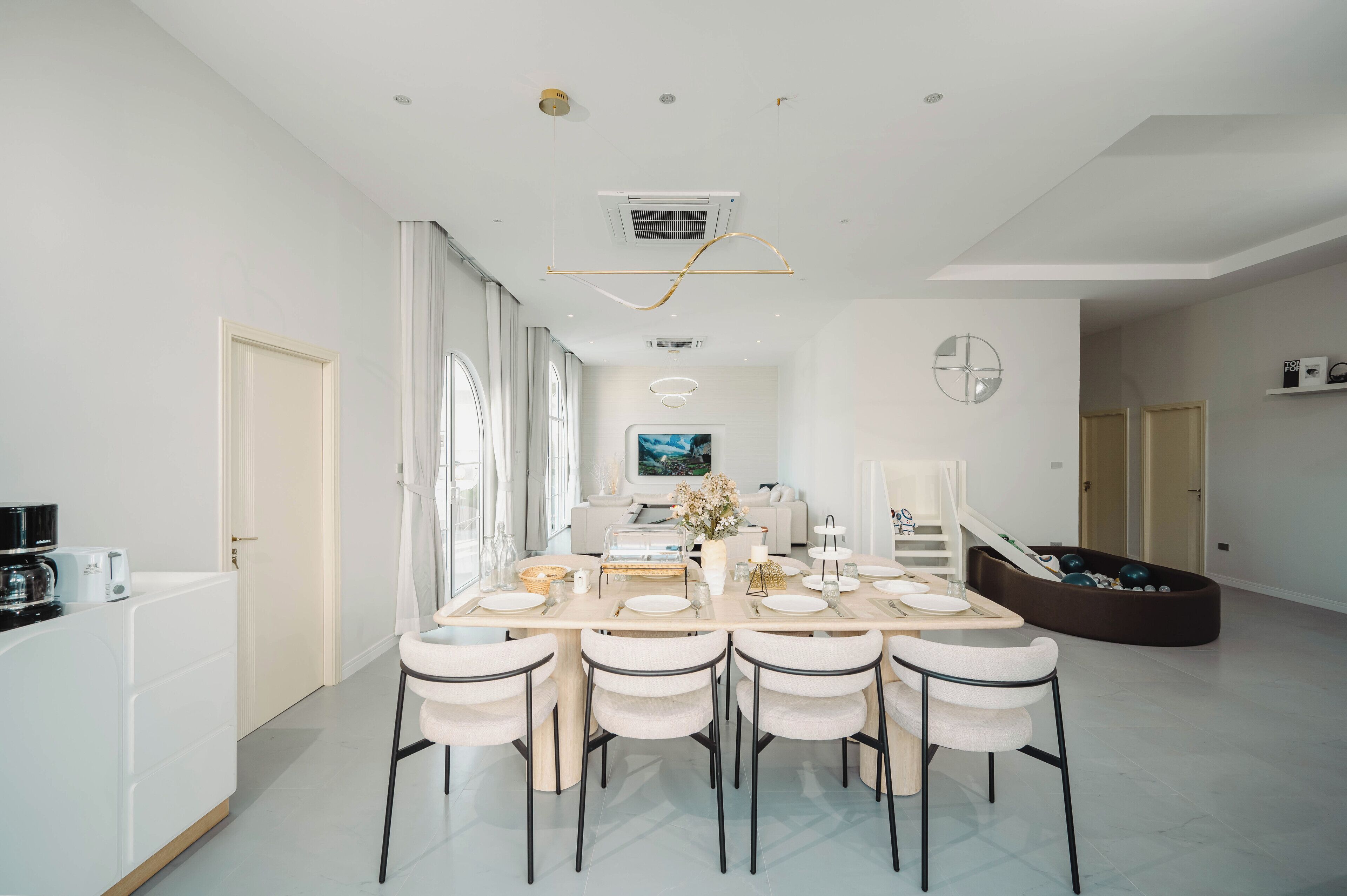 A modern dining area with a large table set for six, surrounded by white chairs. There is a sofa and a television in the background.