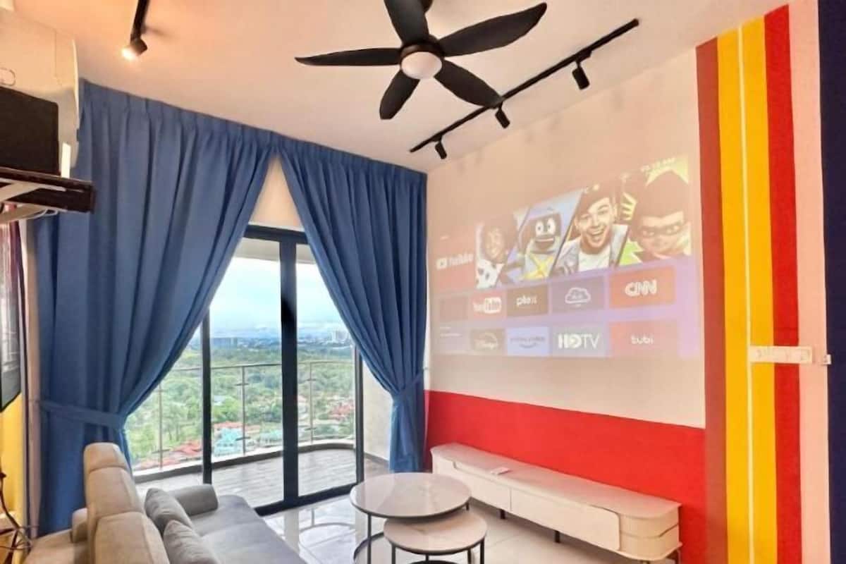 A modern living room with a sofa, a small table, a ceiling fan, a large window with curtains, and a wall with a projection screen displaying various logos.