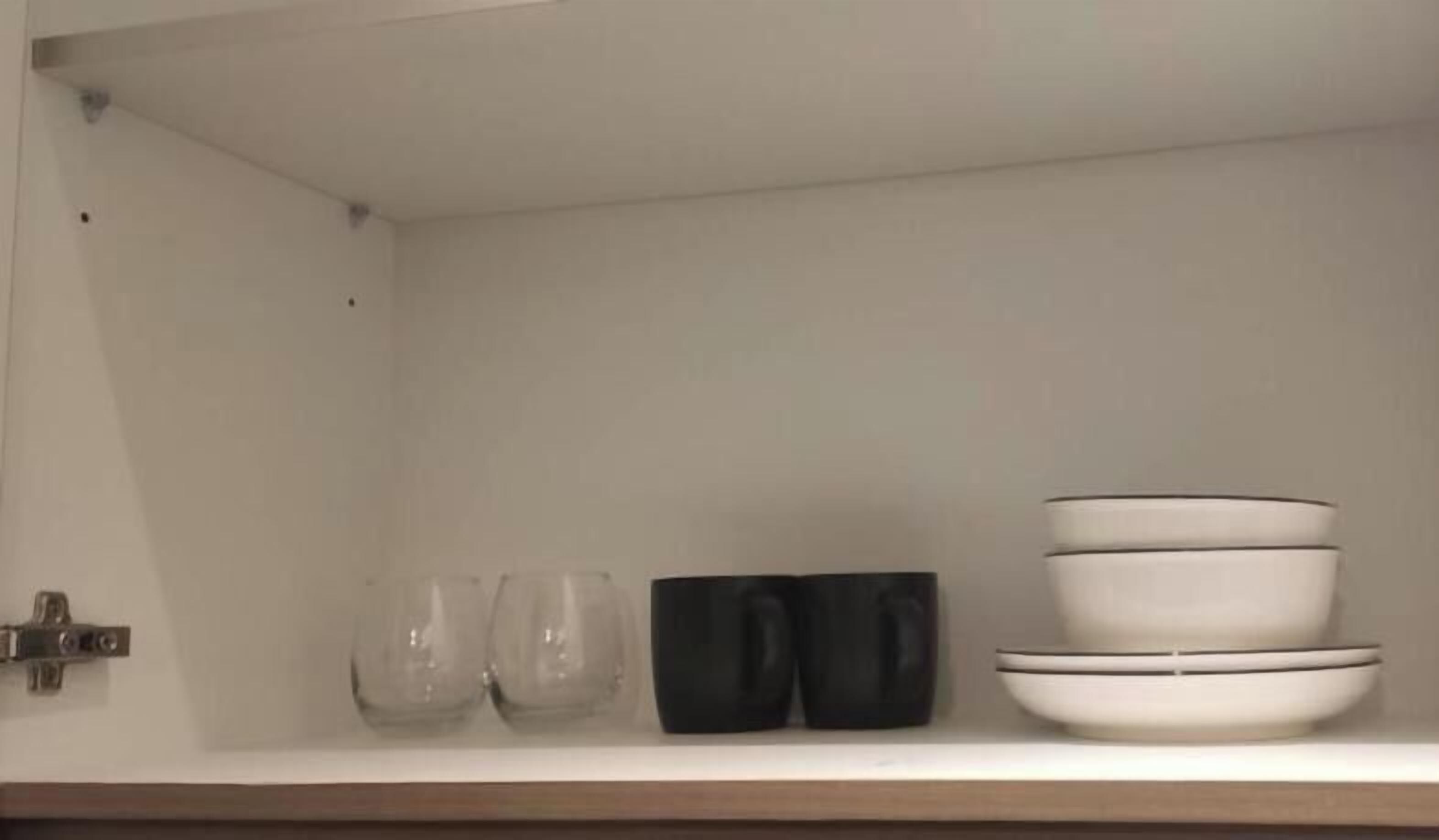 Un armario de cocina con interior blanco, que contiene una pila de cuencos blancos con bordes negros, un recipiente negro y dos vasos transparentes.