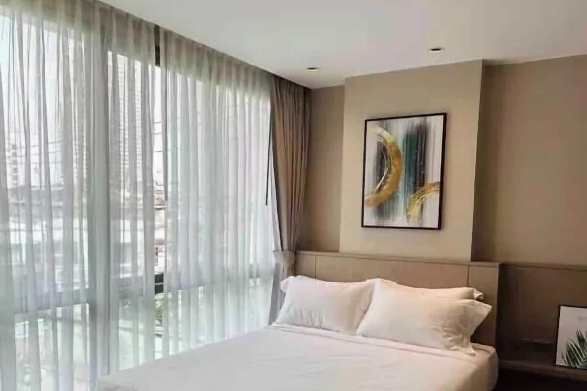 A modern hotel room with a large bed, a view through sheer curtains, a painting on the wall, and a small table.