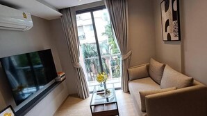 A modern living room with a flat-screen TV, a sofa, a coffee table with a vase, and a balcony view. - Tierra Residence (Bangkok)