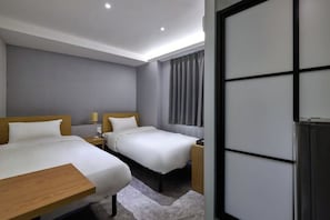 Habitación de hotel con dos camas, una mesita de noche de madera, una pequeña mesa y un ventanal con cortinas.