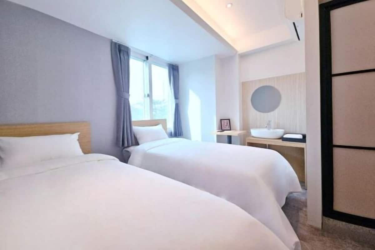 Une chambre d’hôtel avec deux lits, une table de chevet, un miroir rond et une fenêtre avec des rideaux.