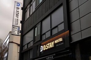 Une enseigne « D.STAY HOSTEL » sur la façade d’un immeuble, avec un motif de fenêtres en réseau.
