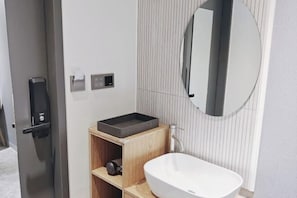 Un baño moderno con un lavamanos blanco, un espejo rectangular y una repisa de madera.