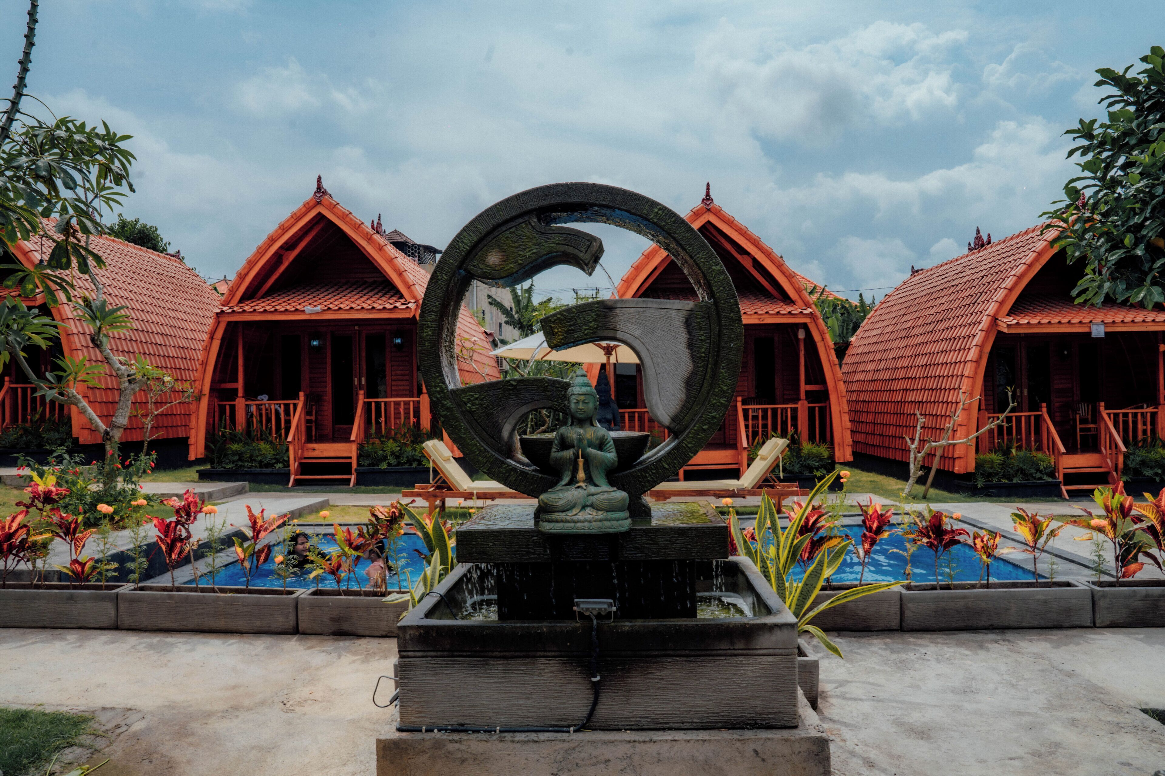A courtyard with a central water feature, surrounded by red-roofed buildings and tropical plants.
