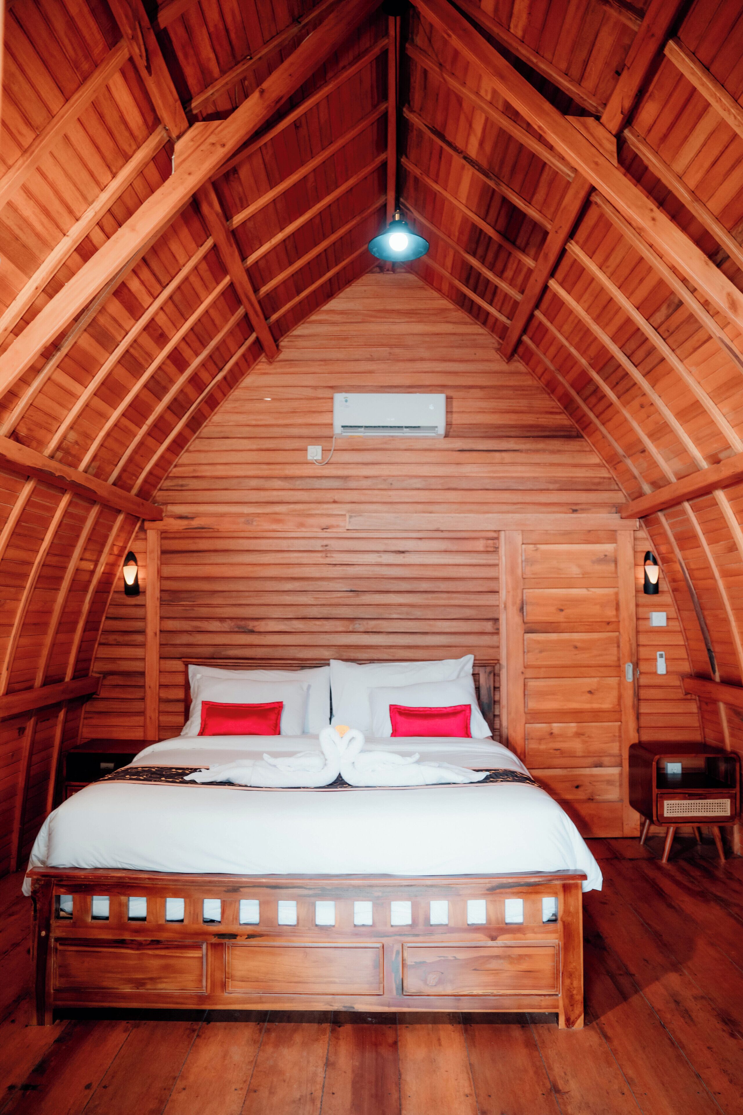A wooden bedroom with a sloped ceiling, a bed with white and red bedding, and a small bedside table.