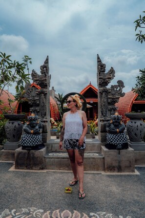 A woman standing in front of a traditional gate with stone statues, a red-roofed building, and tropical plants in the background. - Nusa Dua Huts Pondok Gajah Para by EPS (Nusa Dua)