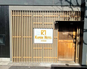 Een hotelentree met een houten poort, een bord waarop 'Kamon Hotel Otemachi' staat en een deur.