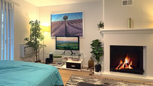 A modern living room with a fireplace, a flat-screen TV mounted on the wall, a bed with a blue bedspread, and a large window with blinds. - Cozy Marina del Rey Stay Near the Beach (Marina del Rey)