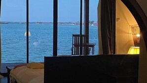 Ένα δω μάτιο με ένα μεγάλο παράθυρο που προσφέρει θέα σε μια έκταση νερού, ένα κρεβάτι, μια καρέκλα και ένα φωτιστικό.