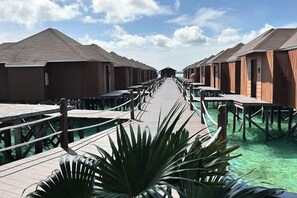 Une passerelle en bois mène à des bungalows sur pilotis, avec une eau turquoise et cristalline de chaque côté.
