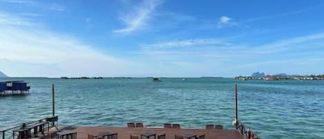 A wooden deck with tables and chairs overlooking calm water, a clear sky, and distant land.