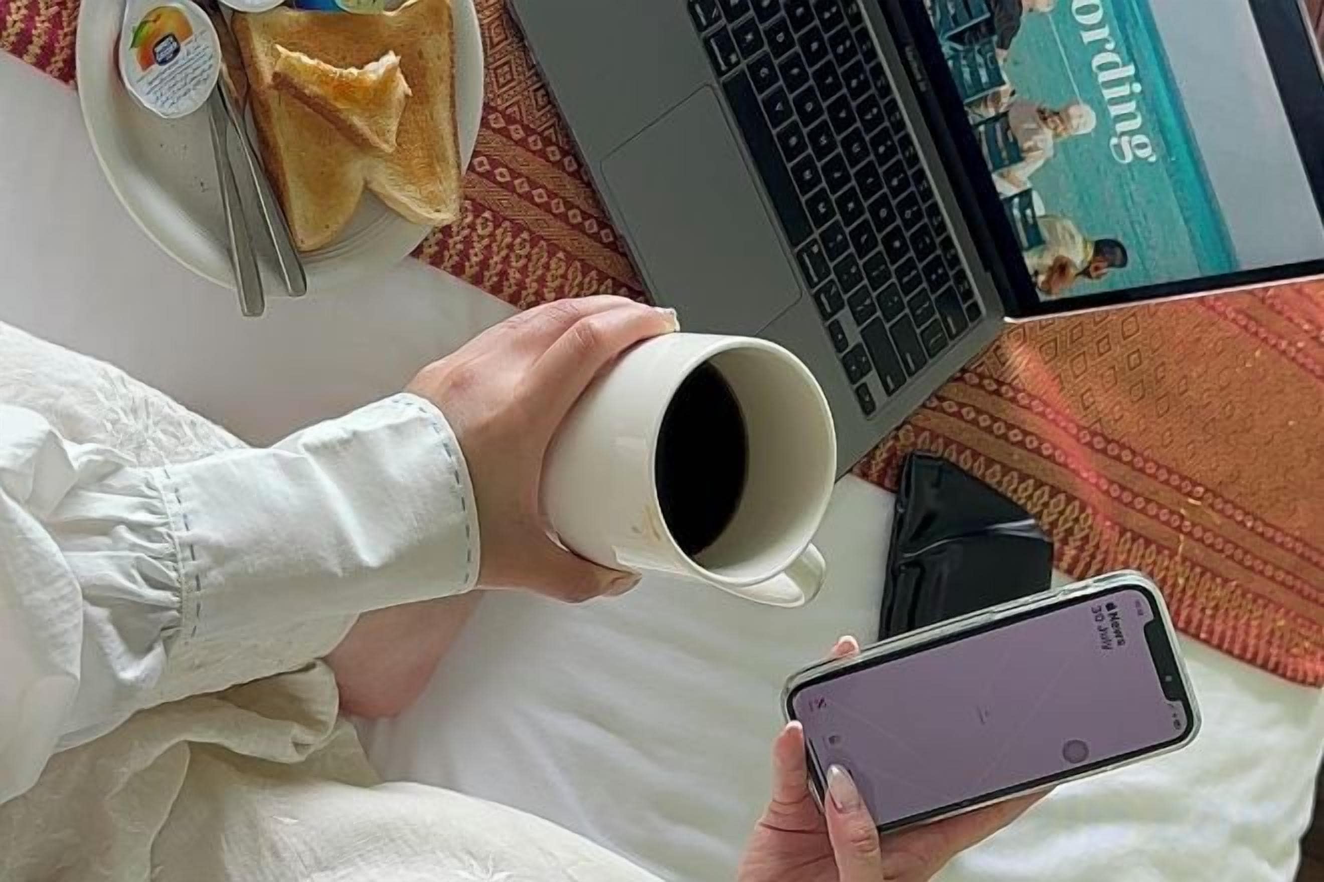 Eine Person hält eine Tasse und ein Smartphone auf einem Bett, auf dem sich ein Laptop und ein Teller mit Essen befinden.