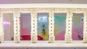 Una stanza con un piano di lavoro in marmo bianco, un grande specchio circondato da composizioni floreali bianche e un murale colorato sullo sfondo.
