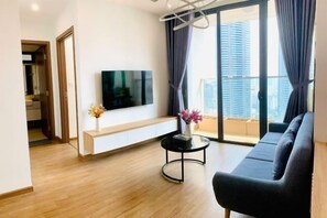 Moderni olohuone, jossa on sohva, valkoiselle tasolle asennettu televisio, pyöreä sohvapöytä ja suuret ikkunat, joista avautuu näkymä kaupunkimaisemaan.