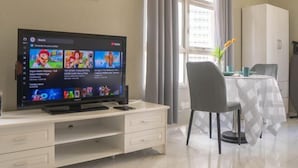 A living room with a flat-screen TV displaying a video game, a white cabinet, a dining table with a white tablecloth, and a window with curtains. - MayTower by Horizon Homes (Kuala Lumpur)