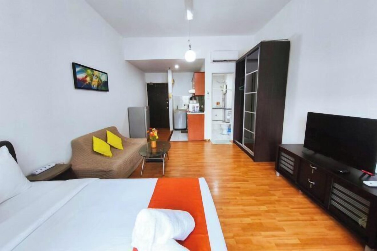 A hotel room with a bed, a sofa, a TV, and a kitchenette in the background.