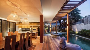 Cocina moderna con armarios de madera y un comedor con sillas de madera. Hay una piscina con una tumbona y una pérgola encima.