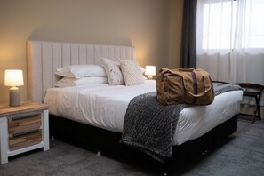 A neatly made bed with pillows, a bedside lamp, a bag, and a chair in a hotel room. - Voltage Lodge (Collie)