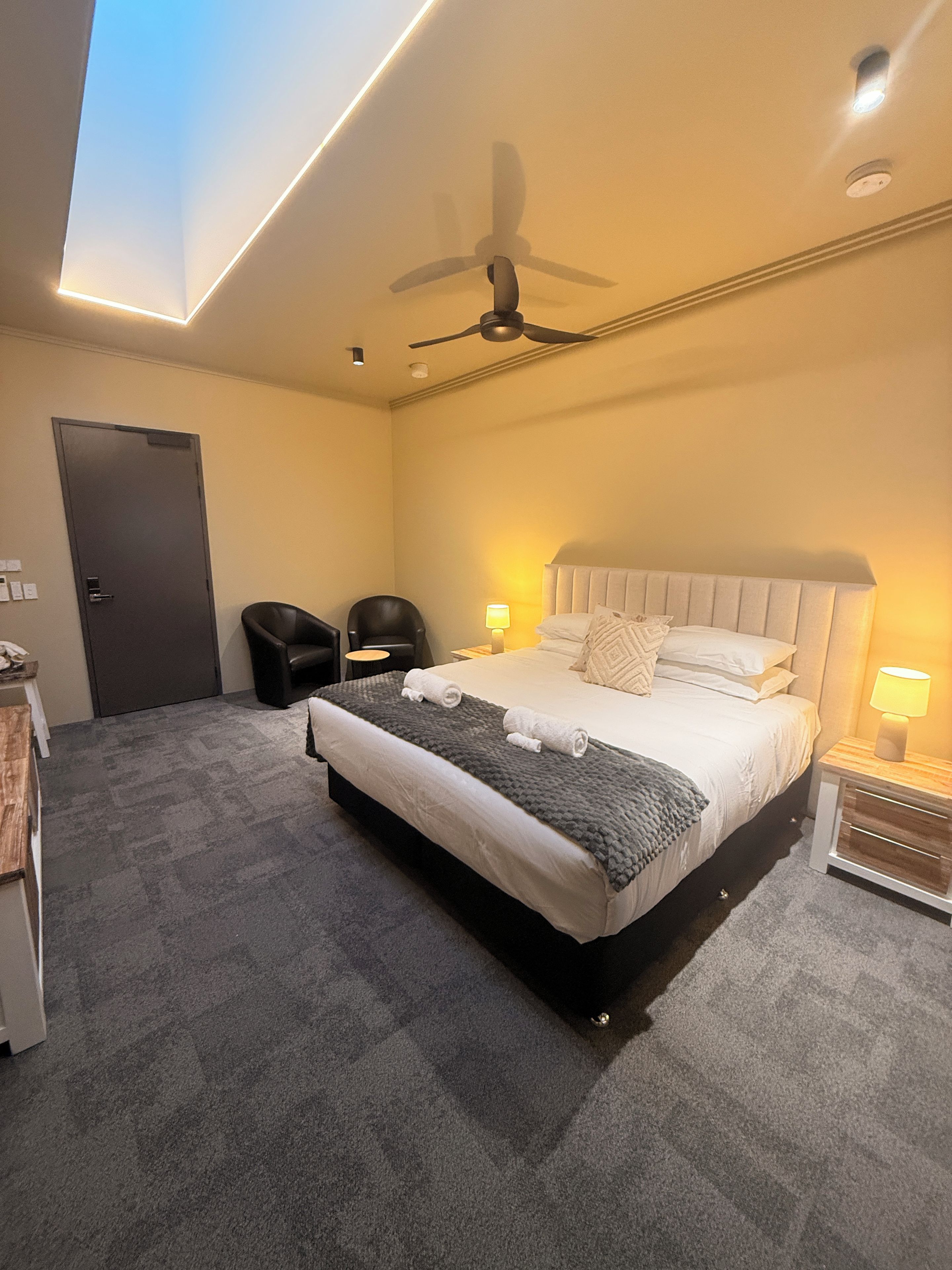 A hotel room with a bed, a ceiling fan, a door, and a small table with a lamp.