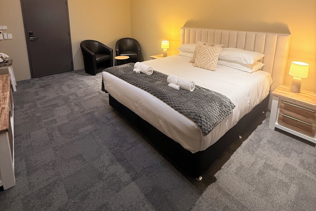 A hotel room with a bed, a ceiling fan, a door, and a small table with a lamp.