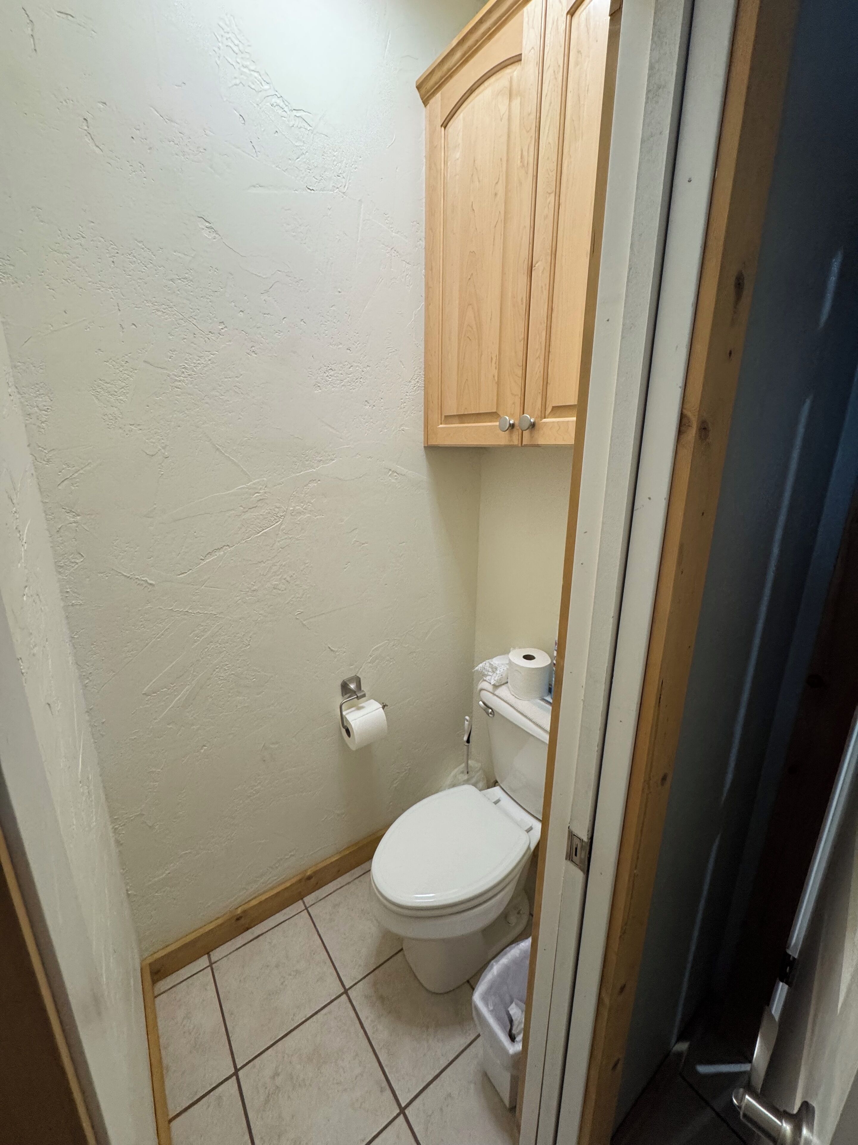 Un baño pequeño con un inodoro, un armario de madera sobre el inodoro y un rollo de papel higiénico en la pared.