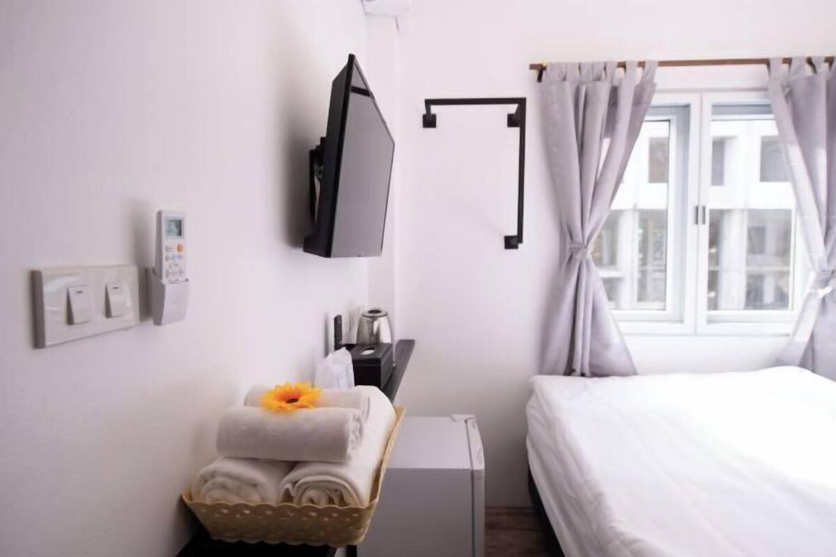 Une chambre d’hôtel avec un lit, un téléviseur fixé au mur, un bureau avec une bouilloire, un panier rempli de serviettes et une fenêtre avec des rideaux.