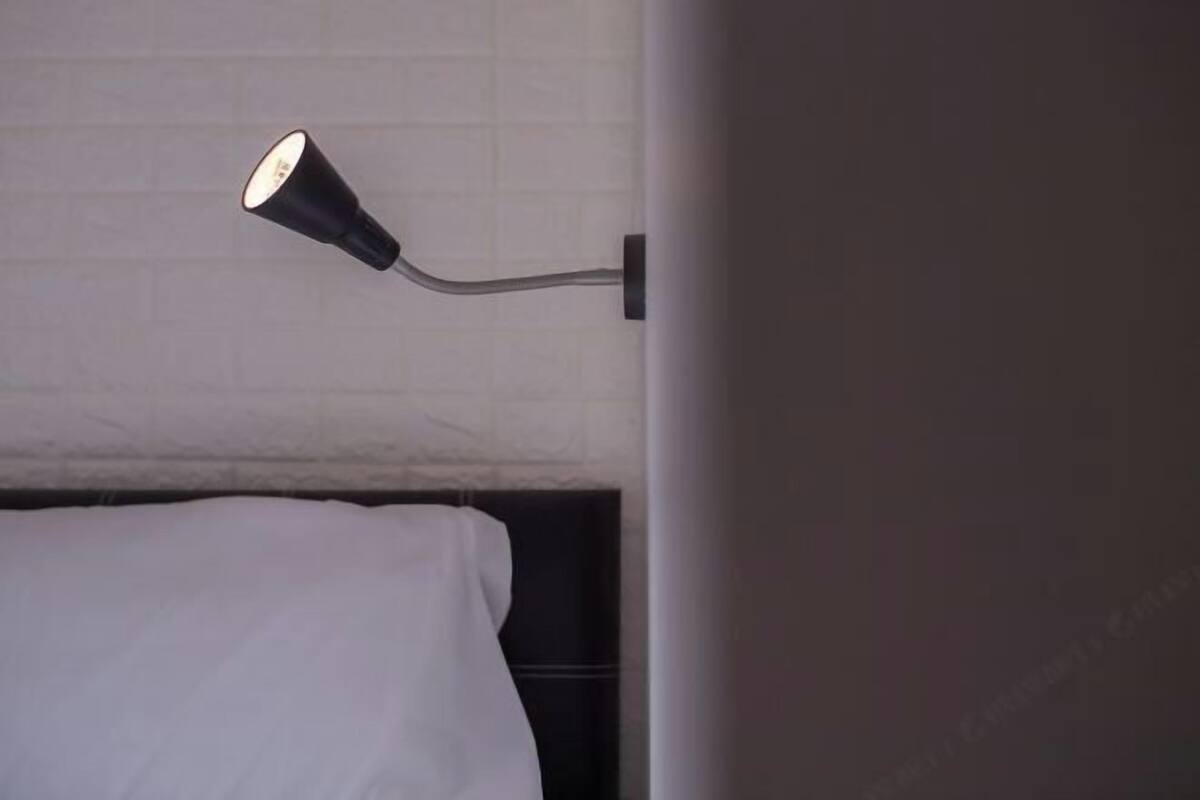 Un lit avec une literie blanche, une lampe noire fixée au mur et un mur recouvert de carreaux blancs.