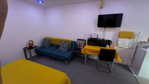 A compact room with a sofa, a small table with a microwave, a chair, a TV mounted on the wall, and a towel rack.
