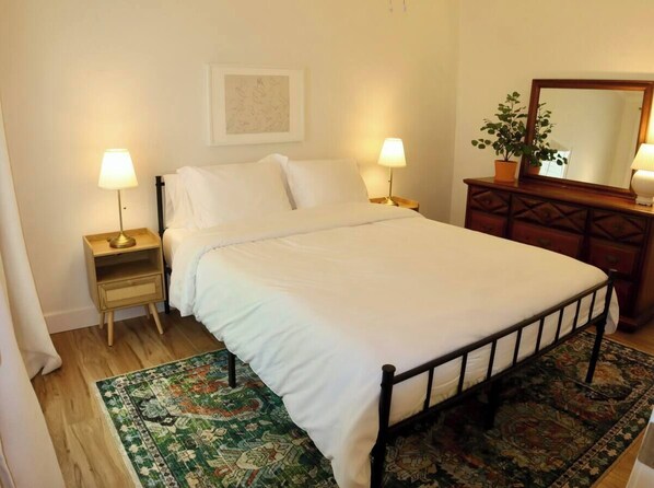 A neatly made bed with white linens, two bedside lamps, a wooden nightstand, a green patterned rug, a wooden dresser with a mirror, and a framed artwork on the wall. - The Gentry Hideaway – A Private Mayberry Retreat (Mount Airy)