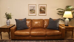 Un sofá de cuero marrón con dos cojines, un tapiz estampado, un jarrón con plantas y dos cuadros en la pared.