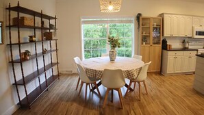 Un comedor con una mesa redonda, cuatro sillas, un jarrón con flores, una estantería de madera y un ventanal con vistas a la vegetación.