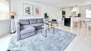 Ein modernes Wohnzimmer mit einem grauen Ecksofa, einem kleinen Couchtisch und einem flauschigen blauen Teppich. Es gibt eine Küchenzeile mit weißen Schränken sowie einen Esstisch mit Stühlen.