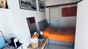 Ein gemütliches Schlafzimmer mit einem Bett, einem Schreibtisch und einem Stuhl. An der Wand hängen Bilder in Rahmen, und es gibt ein Fenster sowie eine Tür.