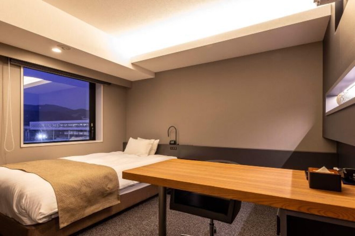 Ein Hotelzimmer mit Bett, Schreibtisch, Fenster mit Blick auf die Stadt und einer Wandlampe.