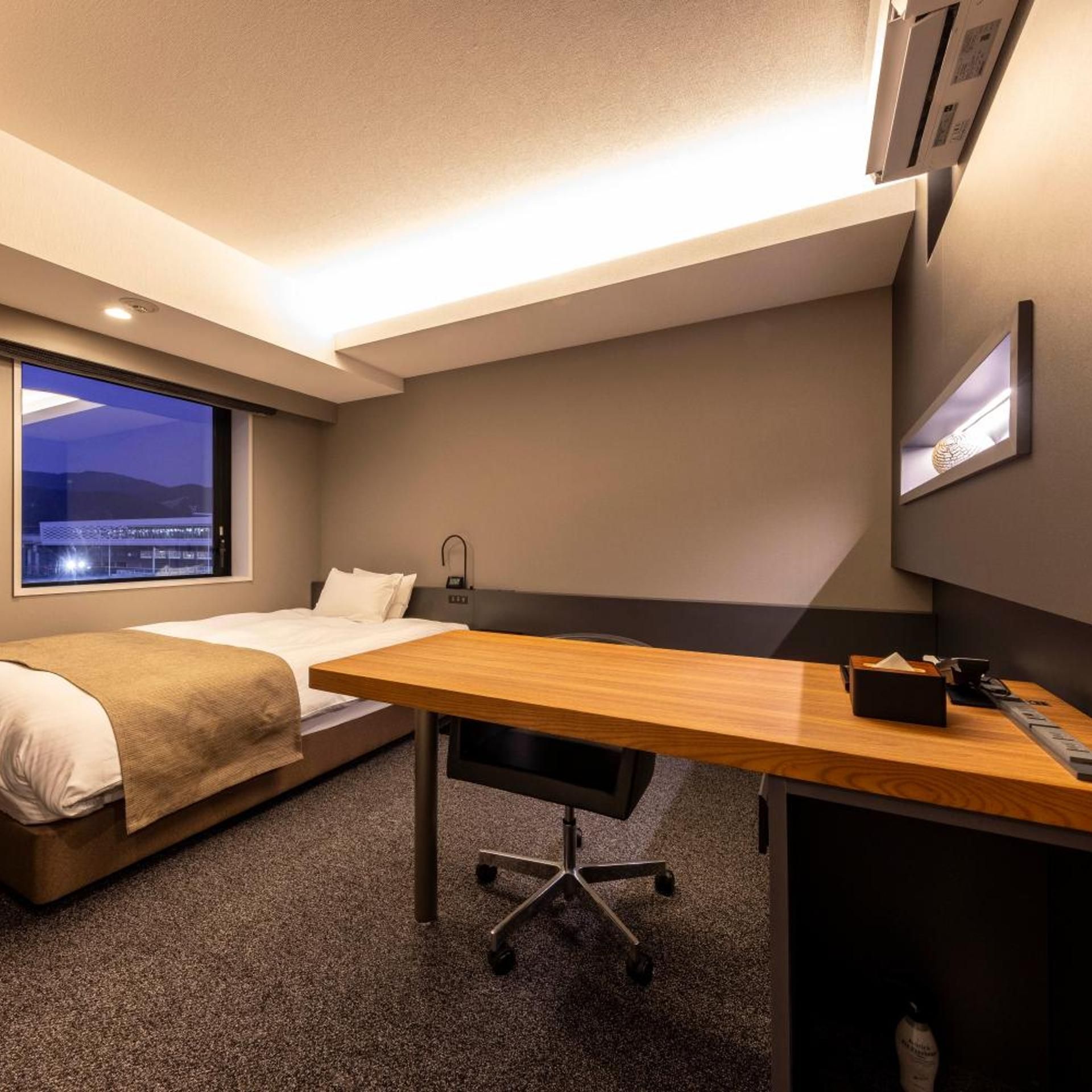 A hotel room with a bed, a desk, and a view of the city at night.