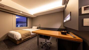A hotel room with a bed, a desk, a chair, a window with a view, and a wall-mounted control panel.
