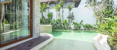 A swimming pool with a wooden deck, surrounded by tropical plants and a stone wall.