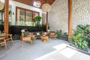 A spacious indoor area with a sofa set, wooden chairs, and a round coffee table. There is a stone wall and a large window providing natural light.