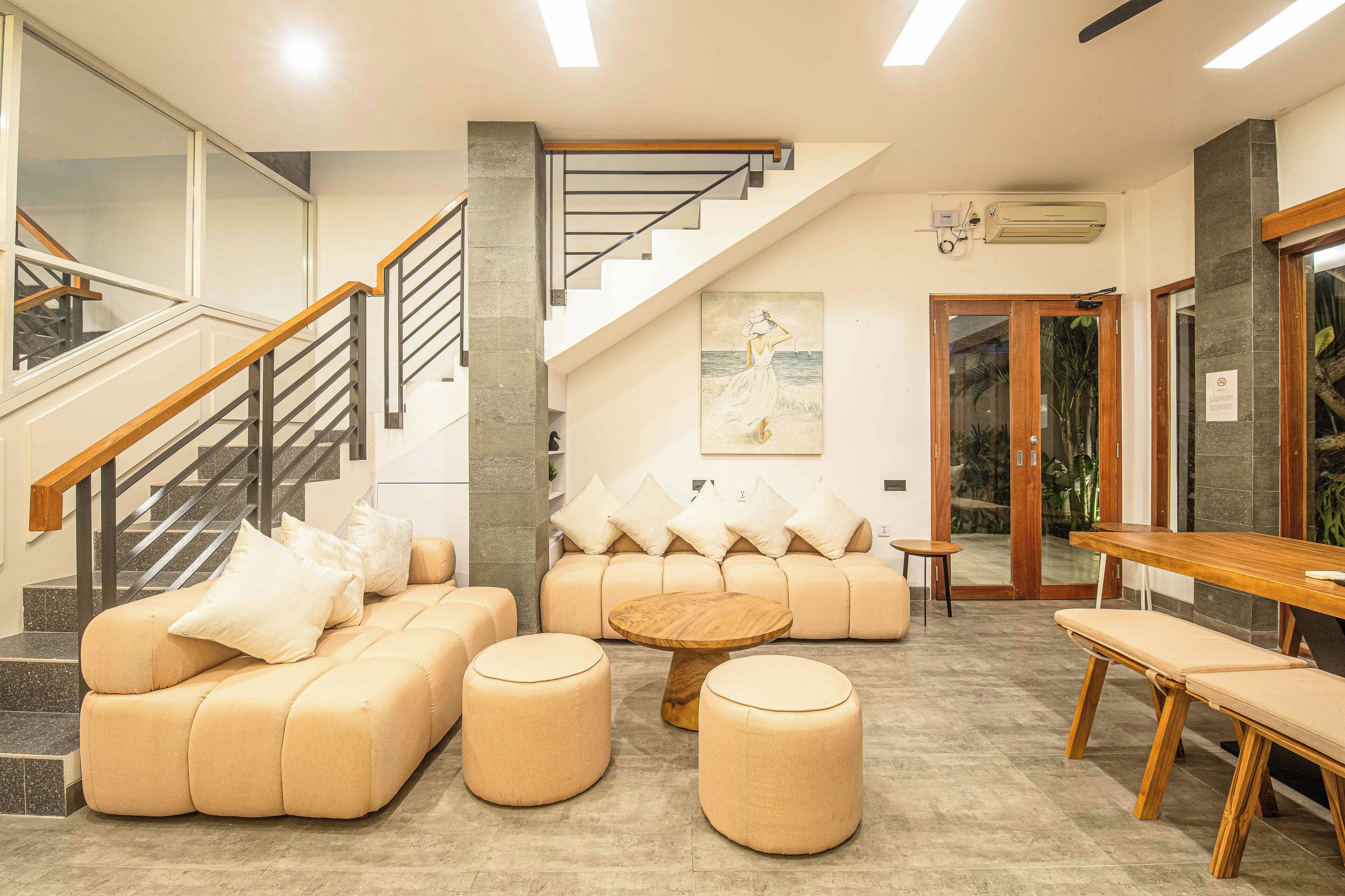 A modern lobby with a staircase, a sofa set, ottomans, a wooden table, and a painting on the wall.