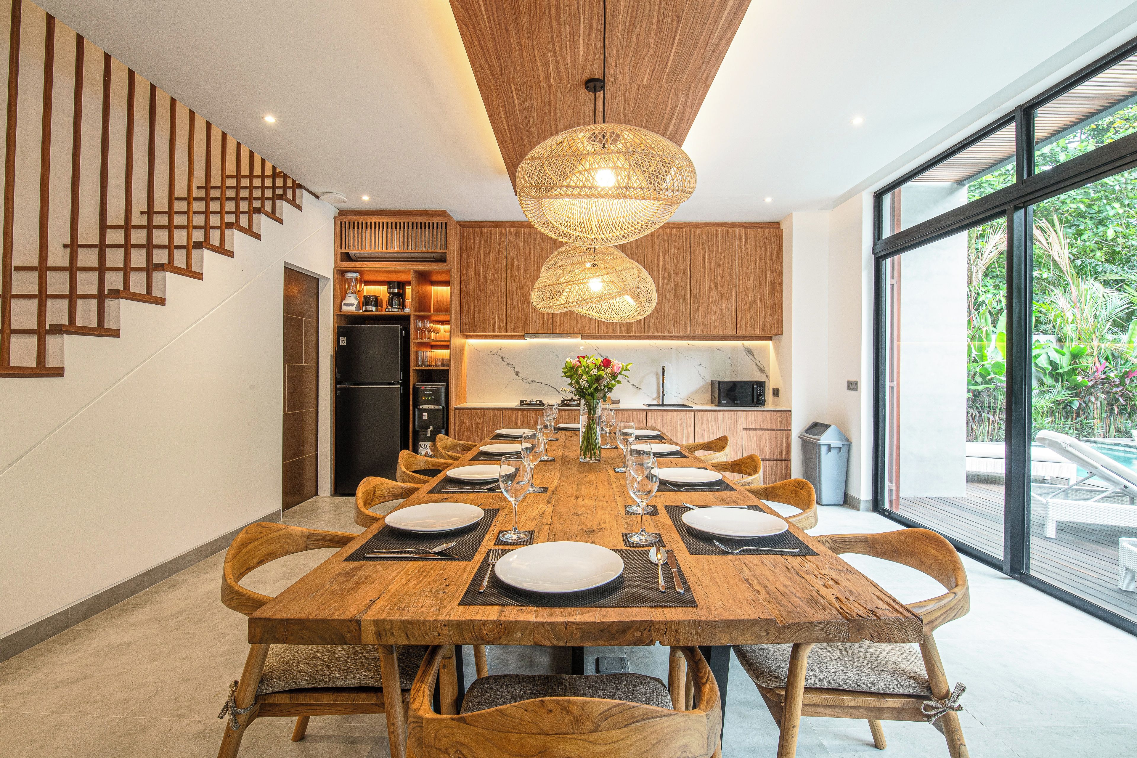 A modern dining area with a wooden table set for six, surrounded by chairs. A staircase with wooden railings is visible to the left.
