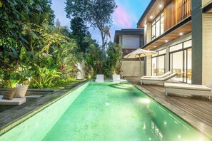 A swimming pool with lounge chairs and a wooden deck, surrounded by lush greenery. - Stylish 5BR Designer Villa, 400m to the Ocean (Seminyak)